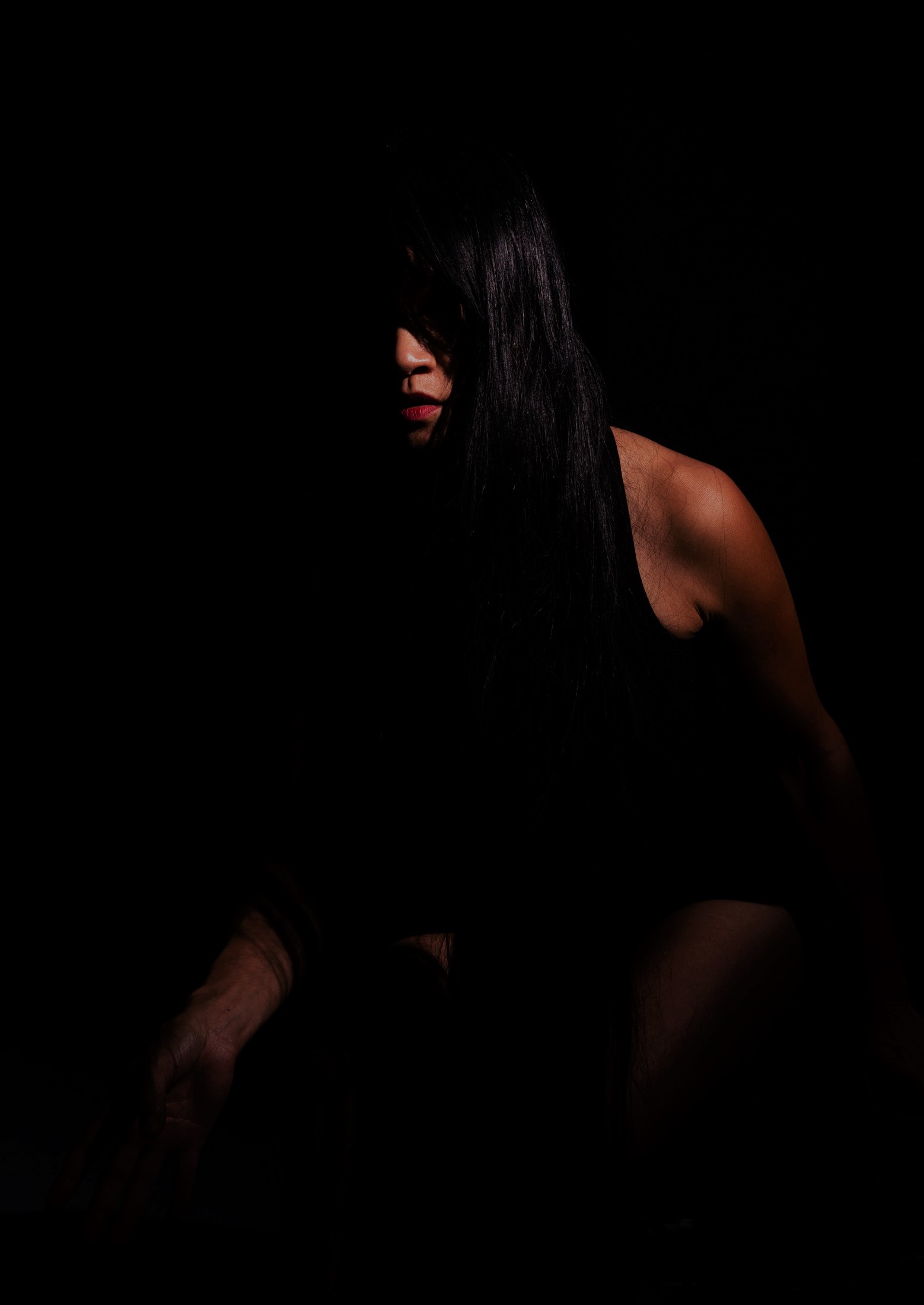Jasmine Orpilla is seated in near-total darkness, partially illuminated by a soft spotlight. Her long black hair frames her face as she leans forward against a black background.