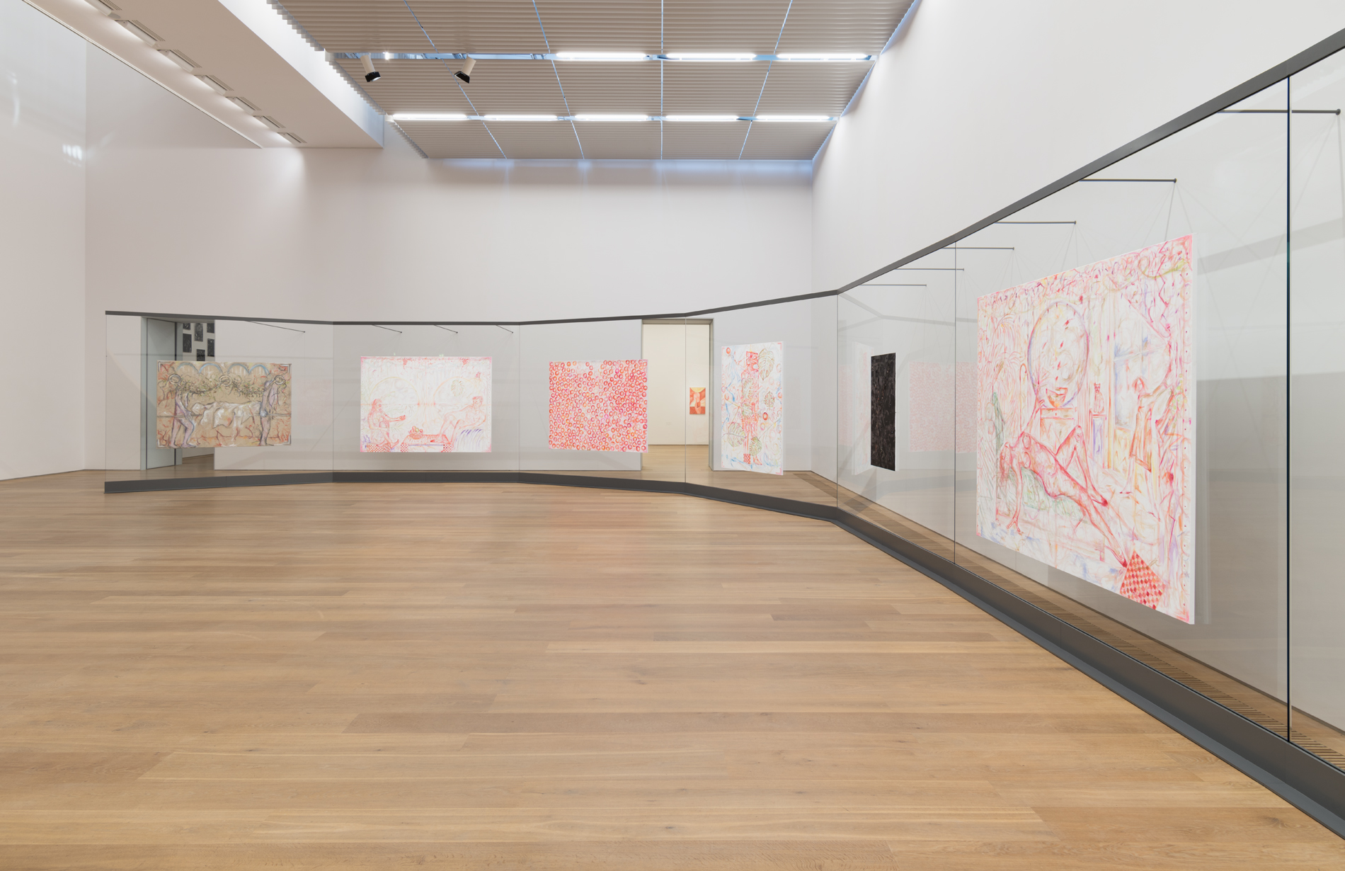 An installation view of multiple paintings displayed along a curved glass wall within a gallery space. The works vary in color and composition, with some featuring abstracted figures and others containing text and intricate linework. 