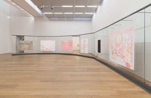 An installation view of multiple paintings displayed along a curved glass wall within a gallery space. The works vary in color and composition, with some featuring abstracted figures and others containing text and intricate linework.