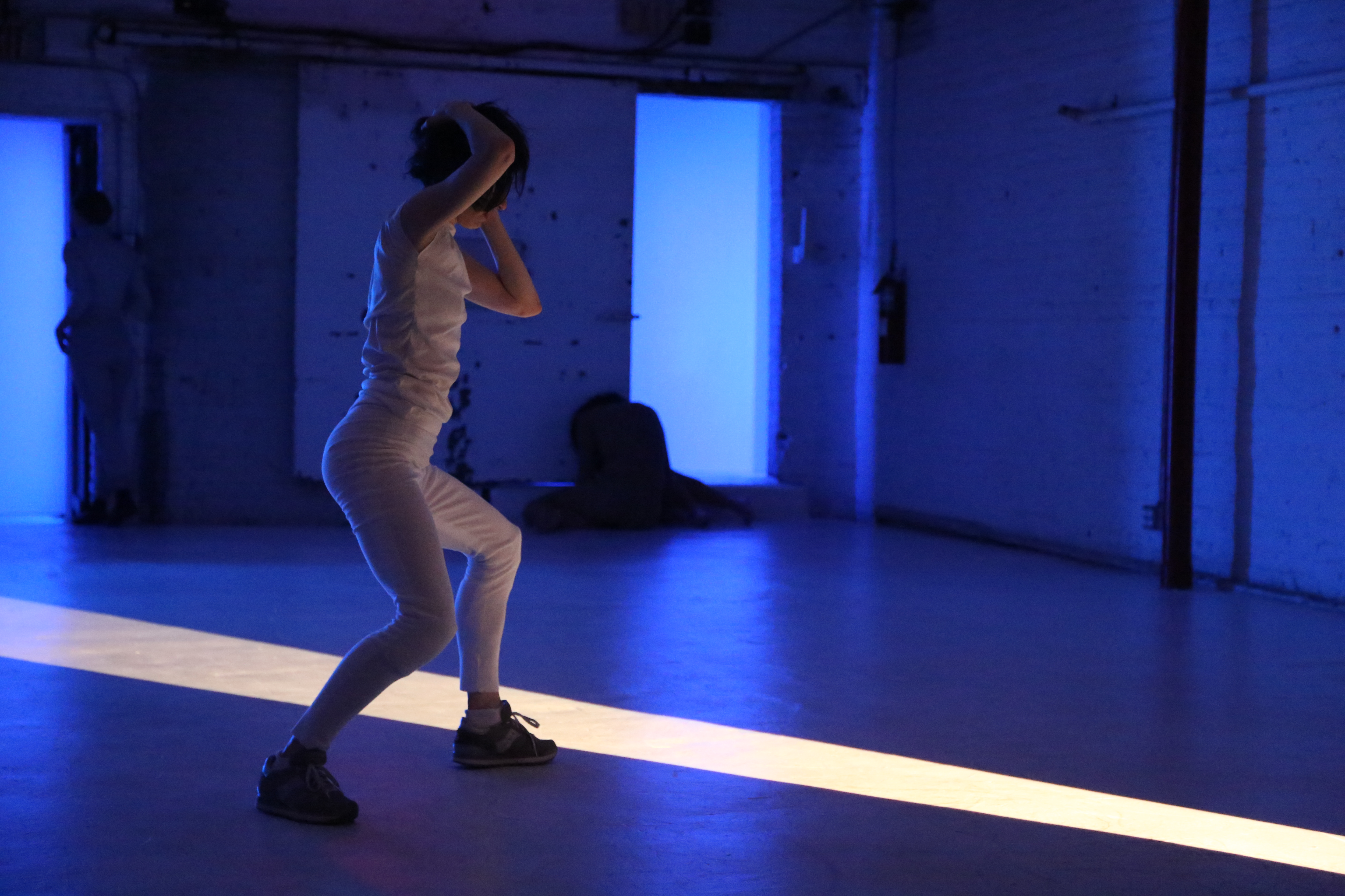 Jennifer Lafferty performs in a dimly lit space wearing an all-white outfit, she is standing and bending her knees slightly, with her hands on her head while turning to the side. A bright strip of light cuts diagonally across the floor, contrasting with the blue lighting that fills the room. A figure is visible in the background, partially obscured by an open doorway.