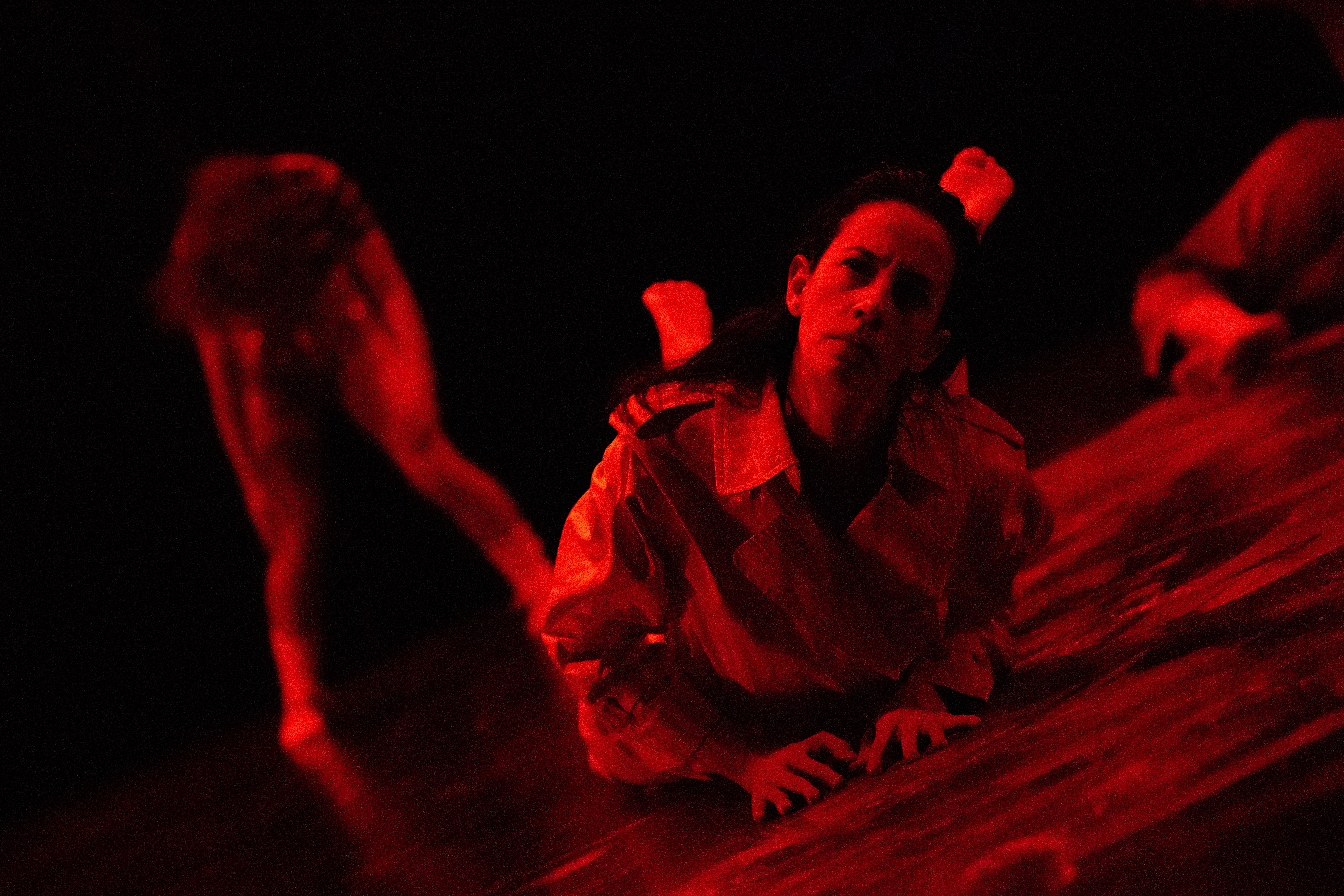 Jennifer Lafferty is shown in red lighting, lying on the floor with her head raised and her hands resting on the ground. She wears a trench coat, and her gaze is tilted to the side. Other performers in sequined outfits are partially visible in the background.