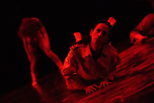 Jennifer Lafferty is shown in red lighting, lying on the floor with her head raised and her hands resting on the ground. She wears a trench coat, and her gaze is tilted to the side. Other performers in sequined outfits are partially visible in the background.