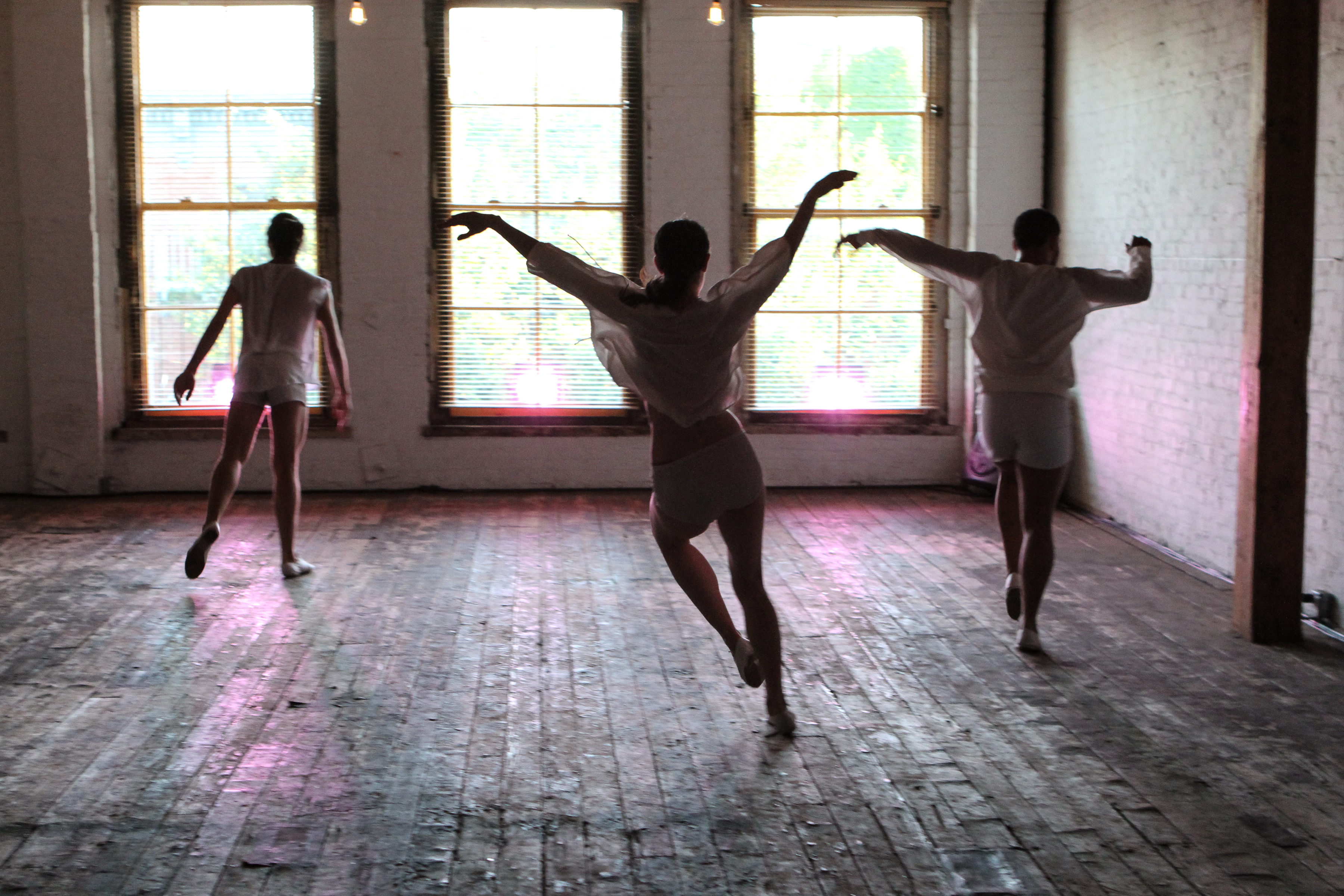 Jennifer Lafferty performs in the center of a wooden floored room, appearing with two other dancers all facing away from the camera in various positions. They all wear sheer white tops and shorts, with their arms extended outward and slightly curved. The image is backlit by large windows, casting the performers in silhouette against the bright natural light streaming in.