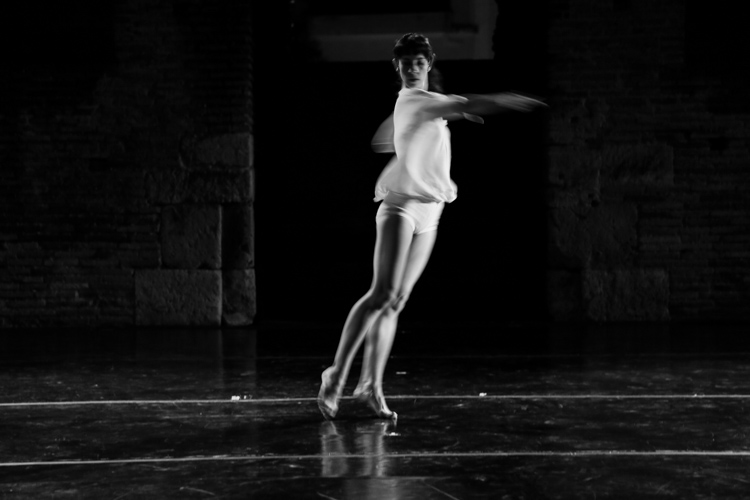 A black and white image of Jennifer Lafferty spinning mid-motion on a stage with a dark, reflective floor. She wears a white top and shorts, with her arms slightly raised and her legs extended. The background features dark, textured stone walls.
