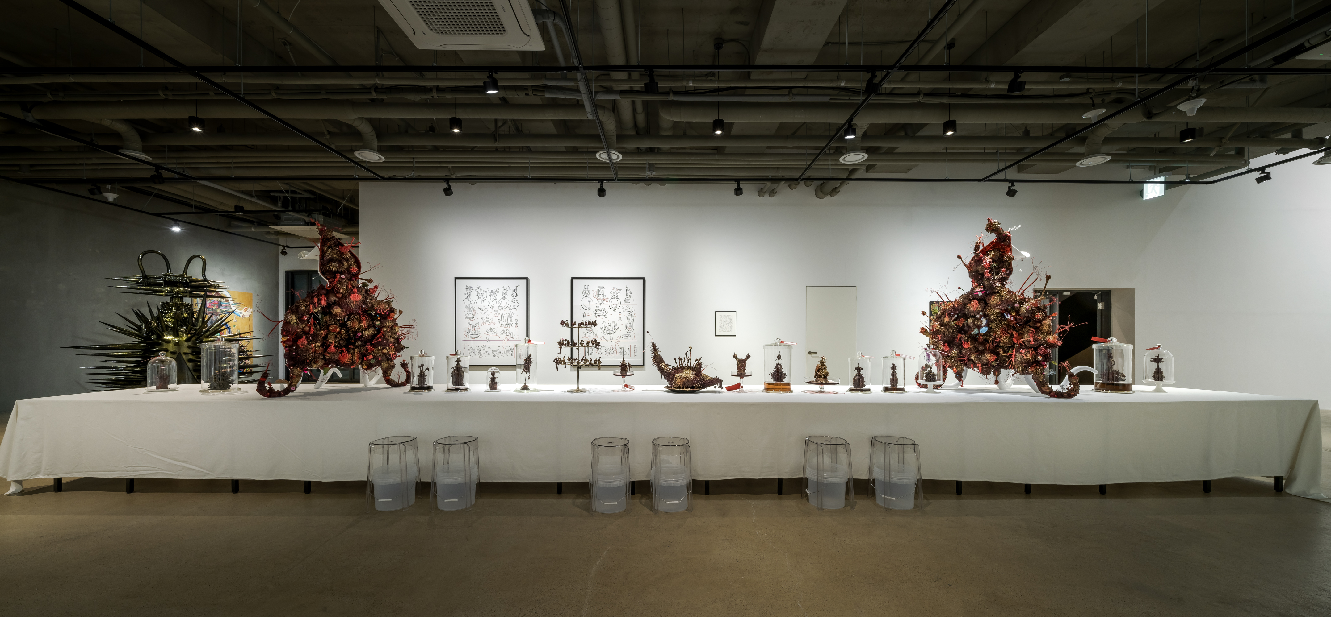 A gallery installation of a long table covered in a white tablecloth containing several spiky bronze-colored statues of creatures in varying sizes displayed on plates and in bell jars. Two larger statues with four tentacles and composed of a dense network of intricate pink, orange, gold, and bronze creatures sit at each end of the table. Detailed black and white drawings of similar creatures hang on the wall behind the table. To the left of the table is a tall spiked black sculpture with golden hues and horns.