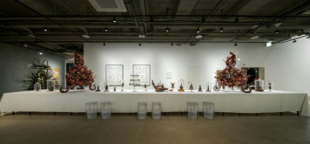 A gallery installation of a long table covered in a white tablecloth containing several spiky bronze-colored statues of creatures in varying sizes displayed on plates and in bell jars. Two larger statues with four tentacles and composed of a dense network of intricate pink, orange, gold, and bronze creatures sit at each end of the table. Detailed black and white drawings of similar creatures hang on the wall behind the table. To the left of the table is a tall spiked black sculpture with golden hues and horns.