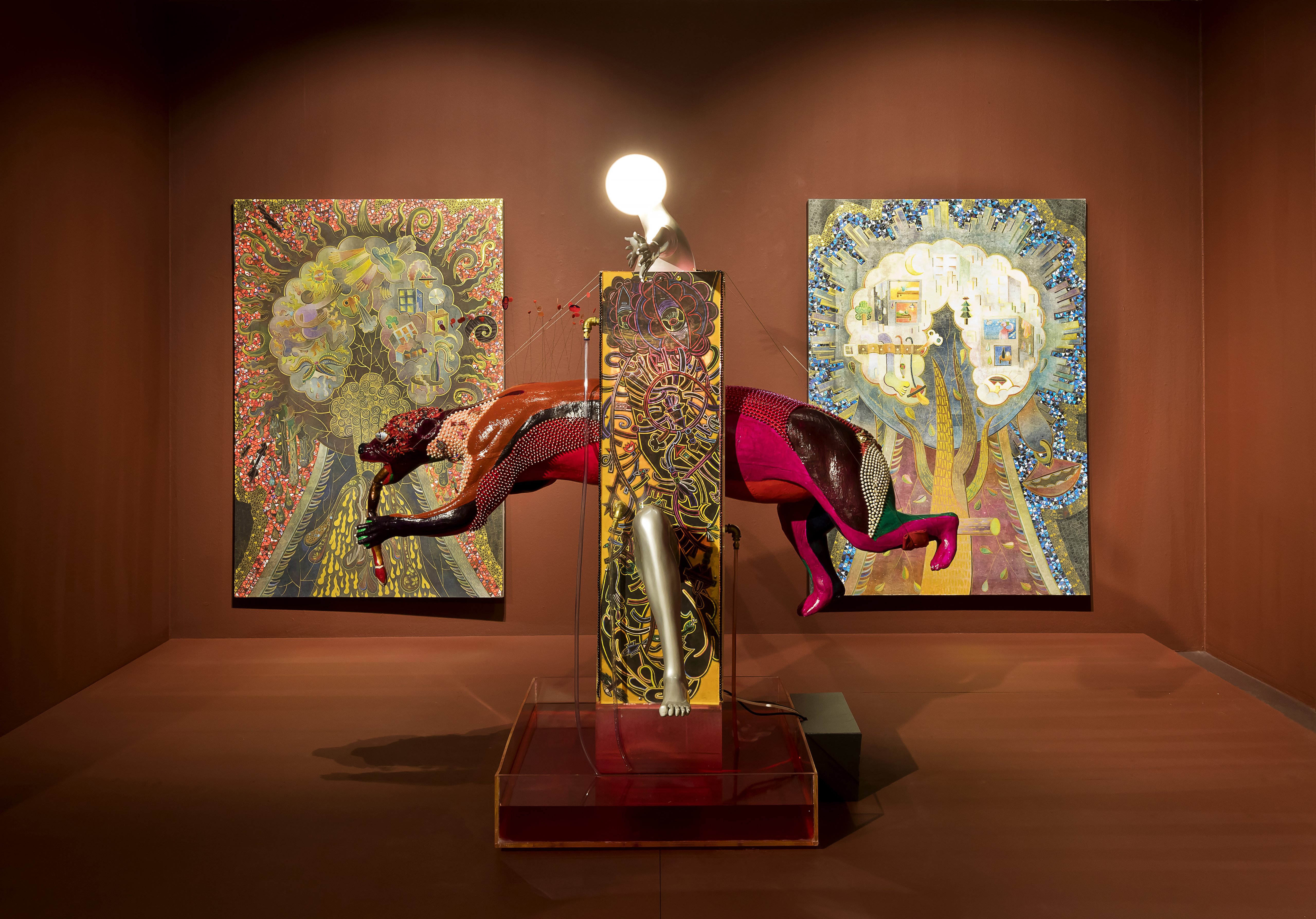 A sculpture of a bear-like creature appearing to run through a wall sits in a gallery space with rust-colored walls and flooring. The sculpture is comprised of a patterned yellow column topped by a lightbulb and the creature is red, pink, orange and partially covered by shiny white beads. Behind the statue are two multicolor abstract patterned paintings of trees with dream sequences superimposed on the crown. 