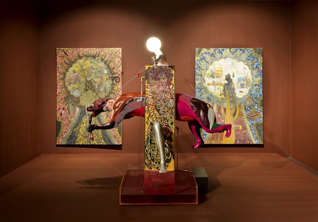 A sculpture of a bear-like creature appearing to run through a wall sits in a gallery space with rust-colored walls and flooring. The sculpture is comprised of a patterned yellow column topped by a lightbulb and the creature is red, pink, orange and partially covered by shiny white beads. Behind the statue are two multicolor abstract patterned paintings of trees with dream sequences superimposed on the crown.