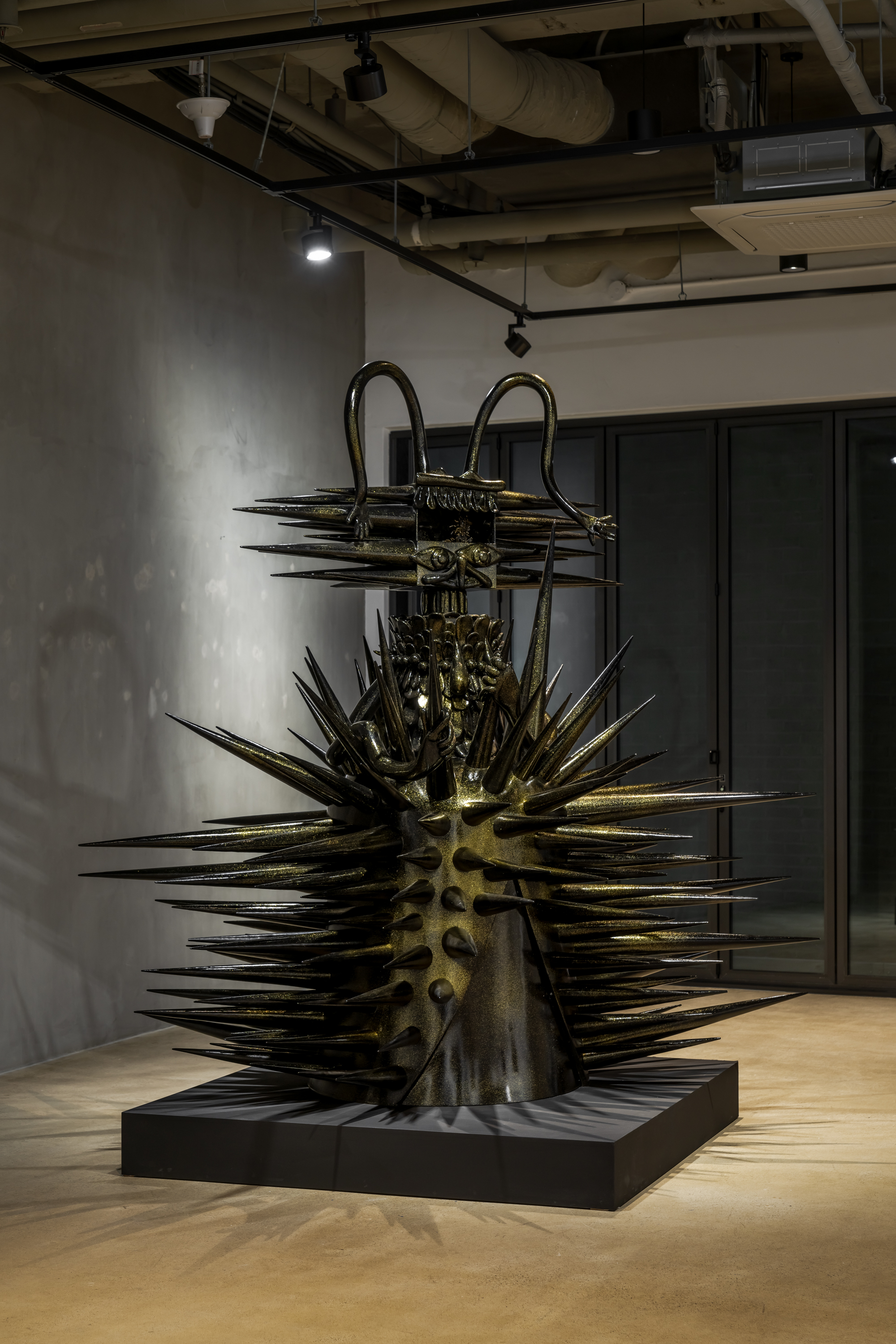 A large black and gold statue of a creature with long spikes protruding from its wide body and head sits on top of a black low pedestal in the gallery space. Shadows of the spikes and the statue’s long antennae are visible on the wall and floor. 