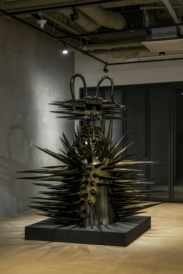 A large black and gold statue of a creature with long spikes protruding from its wide body and head sits on top of a black low pedestal in the gallery space. Shadows of the spikes and the statue’s long antennae are visible on the wall and floor. 