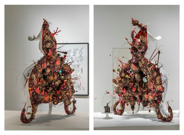 A diptych photograph captures two octopus-like statues seated on a long table covered in a white tablecloth. Each tentacled statue encases a dense cluster of smaller pink, orange, bronze, and gold sculptures of organisms, appearing to mirror one another as though they were bisected down the middle. 