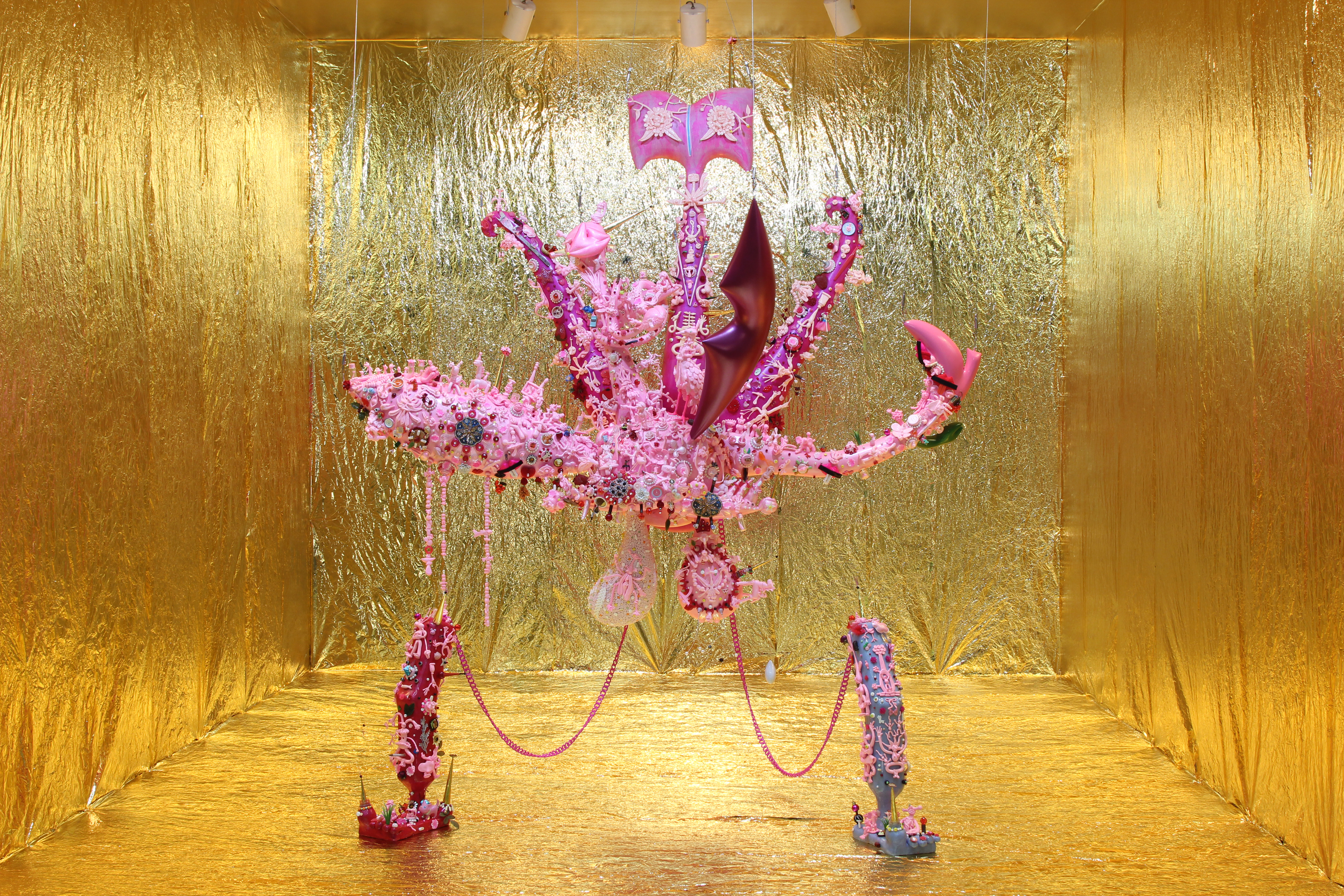 A sculpture of a pink and magenta organism hangs from the ceiling in a gallery space where the walls, floor, and ceiling is covered with metallic gold paper. The sculpture is octopus-like in form, composed of dozens of tightly arranged smaller sculptures of different organisms, and connected by pink chains to a smaller foot-like form anchored to the floor. 