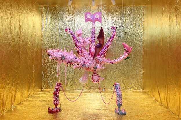 A sculpture of a pink and magenta organism hangs from the ceiling in a gallery space where the walls, floor, and ceiling is covered with metallic gold paper. The sculpture is octopus-like in form, composed of dozens of tightly arranged smaller sculptures of different organisms, and connected by pink chains to a smaller foot-like form anchored to the floor. 