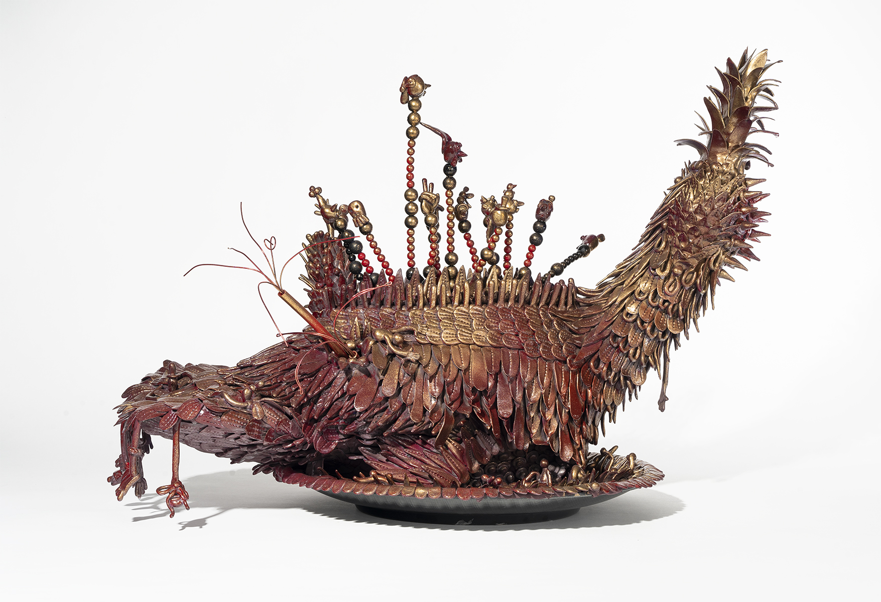 A turkey-like sculpture covered in red and gold metallic feathers rests on a plate composed of the same feathers. Spikes, beaded antennae, and small human-like limbs extend outwards from the creature’s back. 