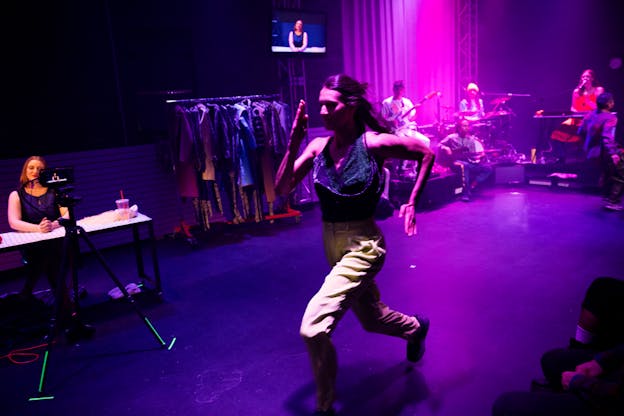 Lena Engelstein runs across a stage lit with purple and pink lights in front of a group of musicians, a clothing rack, and a performer sitting behind a desk being recorded by a camera on a tripod. Lena Engelstein is wearing a sparkly blue top and tan pants and her elbows and knees are pointed at sharp angles.