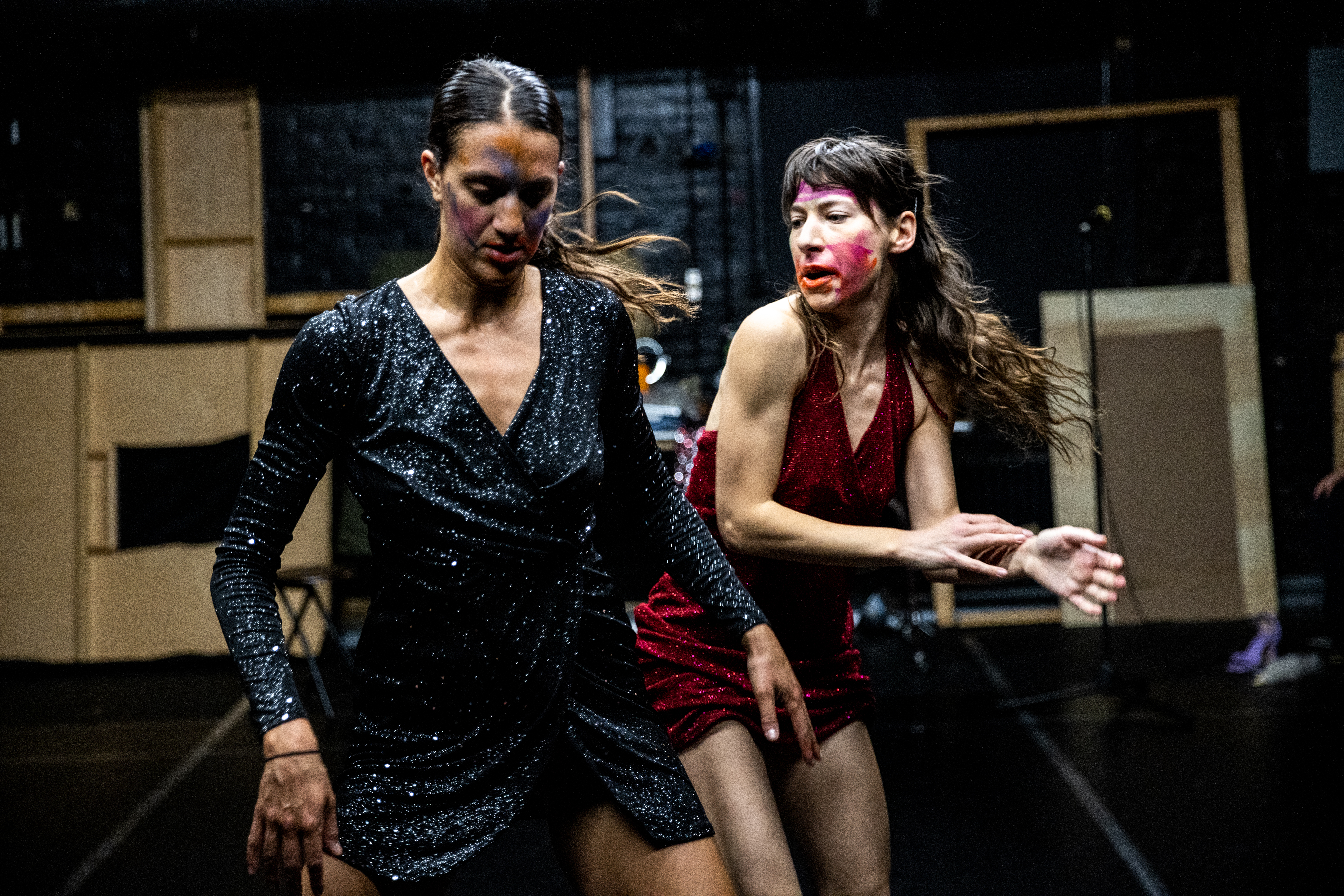 Lena Engelstein and Lisa Fagan, both wearing sparkly dresses, move through a black-walled and floored performance space. Engelstein, wearing blue, looks down and has her arms angled outward. Fagan, positioned behind Engelstein wearing red, looks over her shoulder and gestures with both hands to her right. They both wear streaks of makeup in different colors.