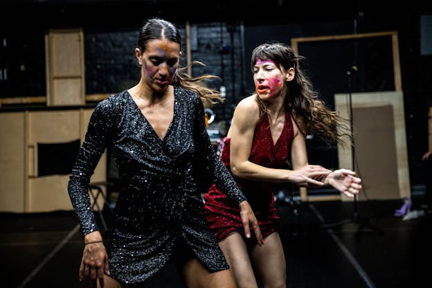 Lena Engelstein and Lisa Fagan, both wearing sparkly dresses, move through a black-walled and floored performance space. Engelstein, wearing blue, looks down and has her arms angled outward. Fagan, positioned behind Engelstein wearing red, looks over her shoulder and gestures with both hands to her right. They both wear streaks of makeup in different colors.