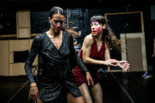 Lena Engelstein and Lisa Fagan, both wearing sparkly dresses, move through a black-walled and floored performance space. Engelstein, wearing blue, looks down and has her arms angled outward. Fagan, positioned behind Engelstein wearing red, looks over her shoulder and gestures with both hands to her right. They both wear streaks of makeup in different colors.