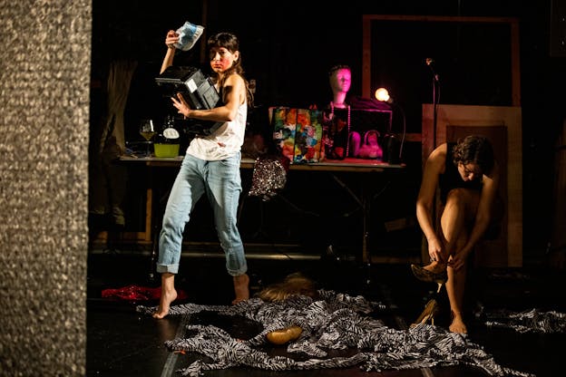 On a stage, Lisa Fagan stands on the left holding a black printer and a fabric pouch while Hannah Mitchell, on the right, sits to put on leopard printed high heels. Zebra printed fabric and a blonde wig lay on the floor around them. Behind the performers, a wine glass, a lamp, a jug that reads “XXX”, patterned bags, and other accessories sit on a table.