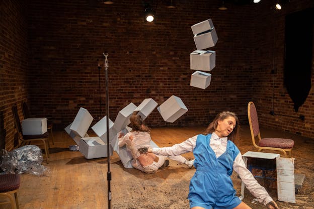 Lena Engelstein and Lisa Fagan are on the floor of a performance space with brick walls and a wooden floor. Engelstein, in the foreground, wears a white shirt under a blue romper. Her arms are outstretched and in one hand she holds dirt. Fagan, wearing a white shirt and pants, is sitting on the background facing away from the audience while she grabs one of several grey boxes that are toppling over. There are several wooden chairs with red seat cushions scattered throughout the scene.