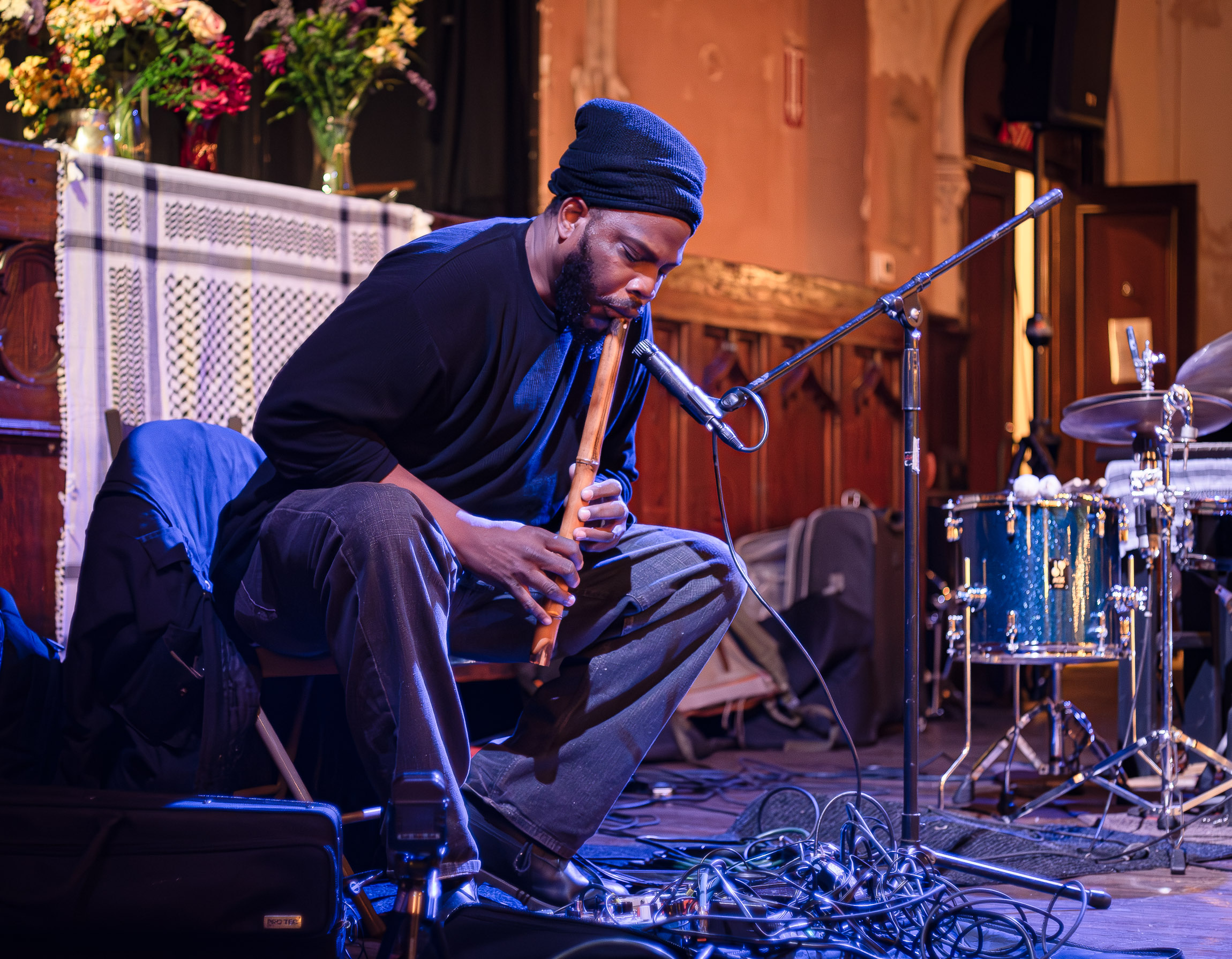 Jamal Moore is seated playing a wooden flute into a microphone while wearing a black beanie and dark clothing. He leans slightly forward with his eyes closed and hands positioned on the instrument as he plays and his eyes closed. The setting is an indoor performance space with flowers arranged in clear vases sitting on a ledge and a patterned scarf hanging in the background. To his side sits a drum kit and at his feet are several tangled musical cables.
