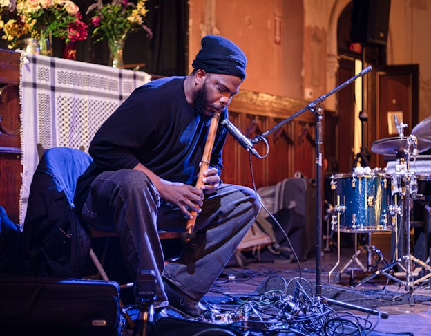 Jamal Moore is seated playing a wooden flute into a microphone while wearing a black beanie and dark clothing. He leans slightly forward with his eyes closed and hands positioned on the instrument as he plays and his eyes closed. The setting is an indoor performance space with flowers arranged in clear vases sitting on a ledge and a patterned scarf hanging in the background. To his side sits a drum kit and at his feet are several tangled musical cables.