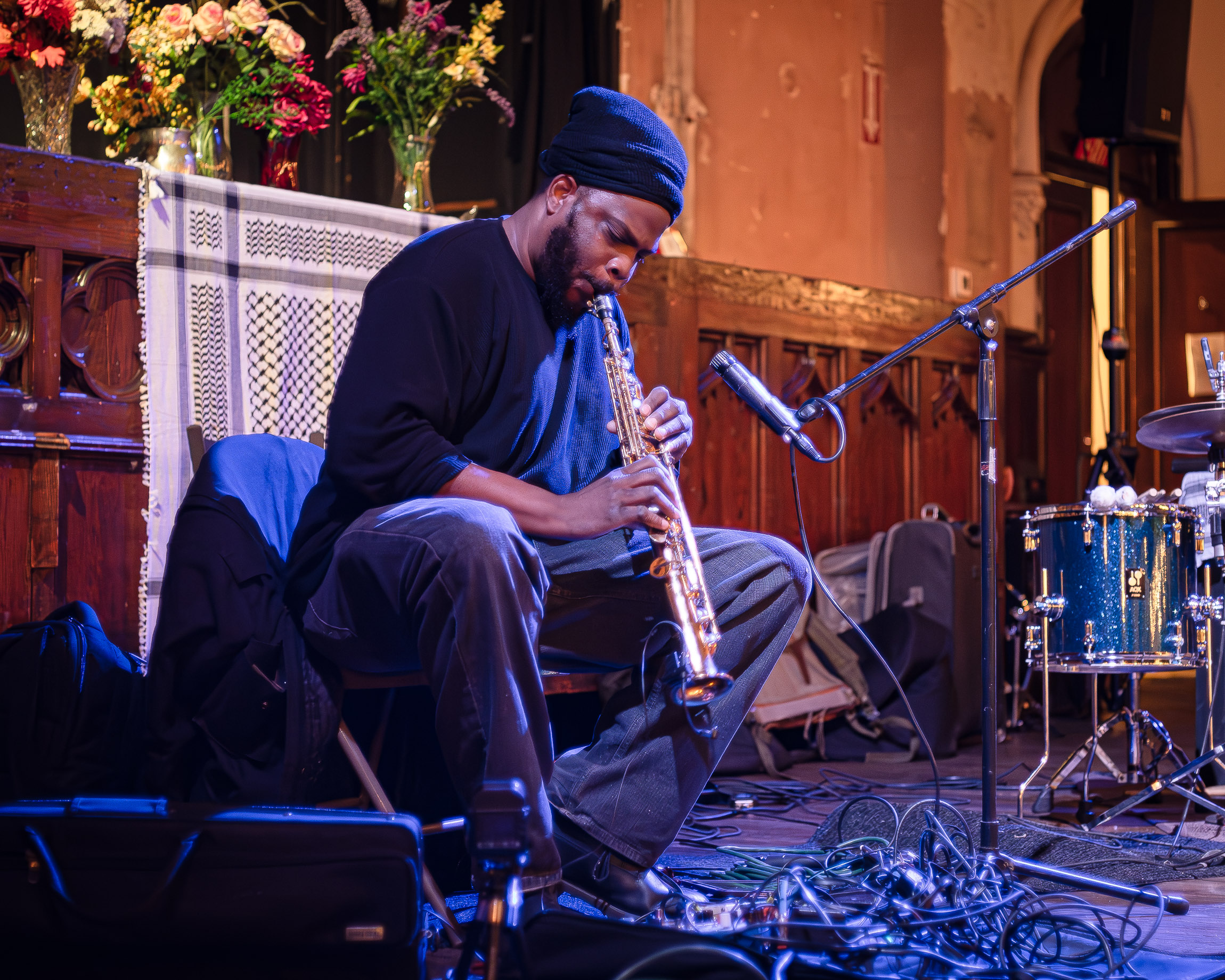 Jamal Moore is seated playing a soprano saxophone while wearing a black beanie and dark clothing. His hands grip the instrument while his lips press against the mouthpiece. A microphone stand is positioned in front of h im and to side is a drum kit. Behind Jamal is a wooden ledge adorned with flowers arranged in clear vases. A patterned scar f hangs from the ledge and at his feet are several tangled musical cables
