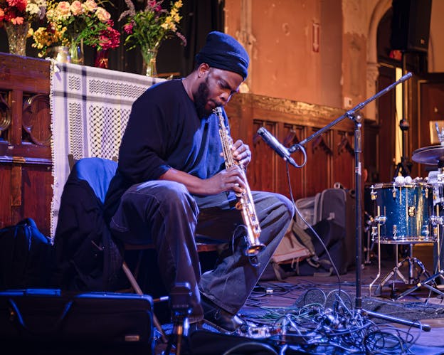 Jamal Moore is seated playing a soprano saxophone while wearing a black beanie and dark clothing. His hands grip the instrument while his lips press against the mouthpiece. A microphone stand is positioned in front of h im and to side is a drum kit. Behind Jamal is a wooden ledge adorned with flowers arranged in clear vases. A patterned scar f hangs from the ledge and at his feet are several tangled musical cables