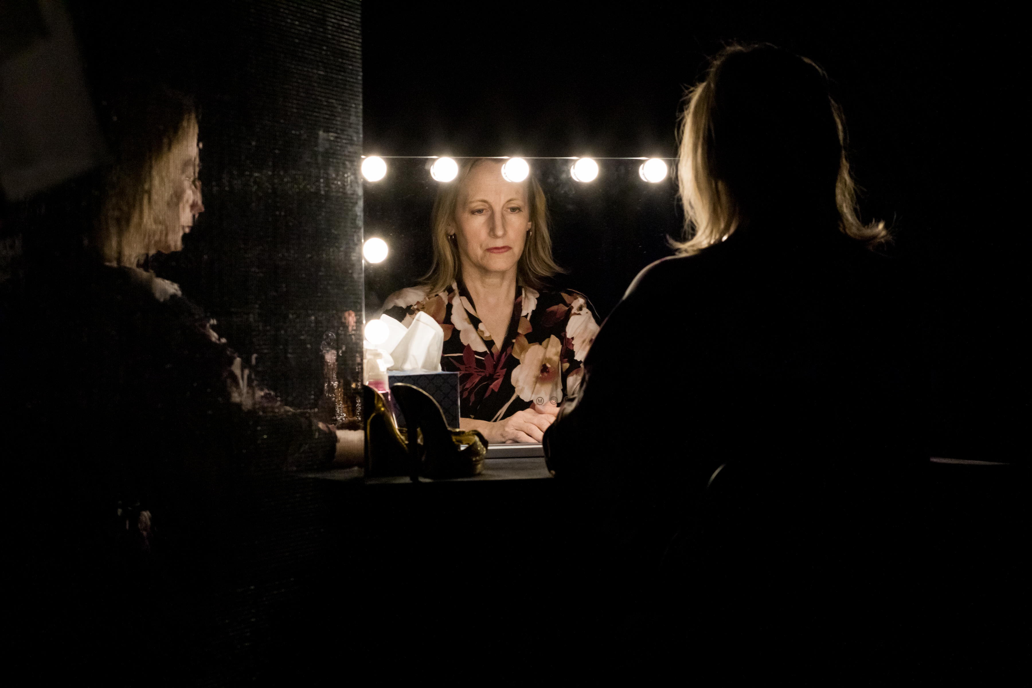 Peggy Piacenza gazes into her well-lit reflection in a dressing room mirror within a dark room. She wears a floral robe. To her left is another pixelated reflection of the side of her face and body. A box of tissues sits in front of the mirror. The background and foreground are both dark/in shadow.