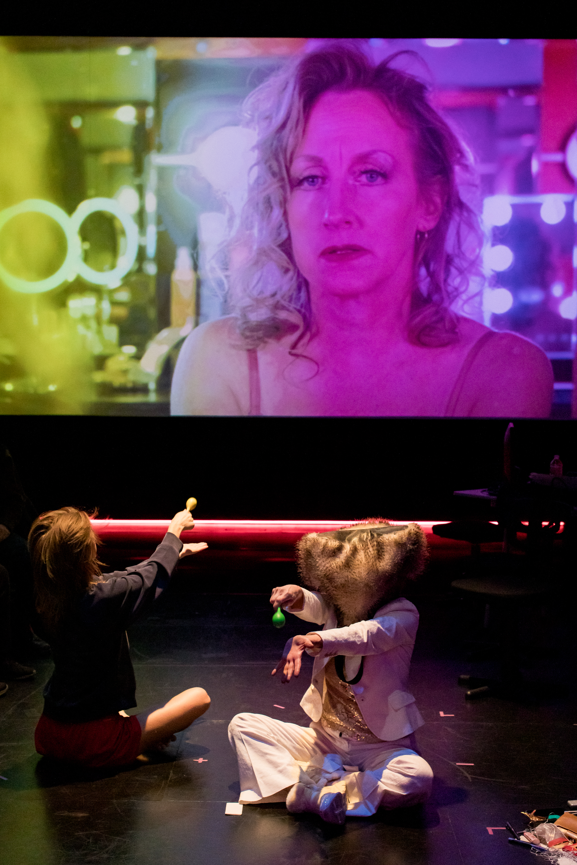 Two performers sit cross legged on a stage holding their arms out beneath a large projected image of Peggy Piacenza who stares into the camera bathed in green, blue, and purple light. One performer faces the audience and wears a white suit with a gold vest and a fur hat covering their head. The other performer, facing the image of Piacenza, wears a black suit jacket and red shorts.