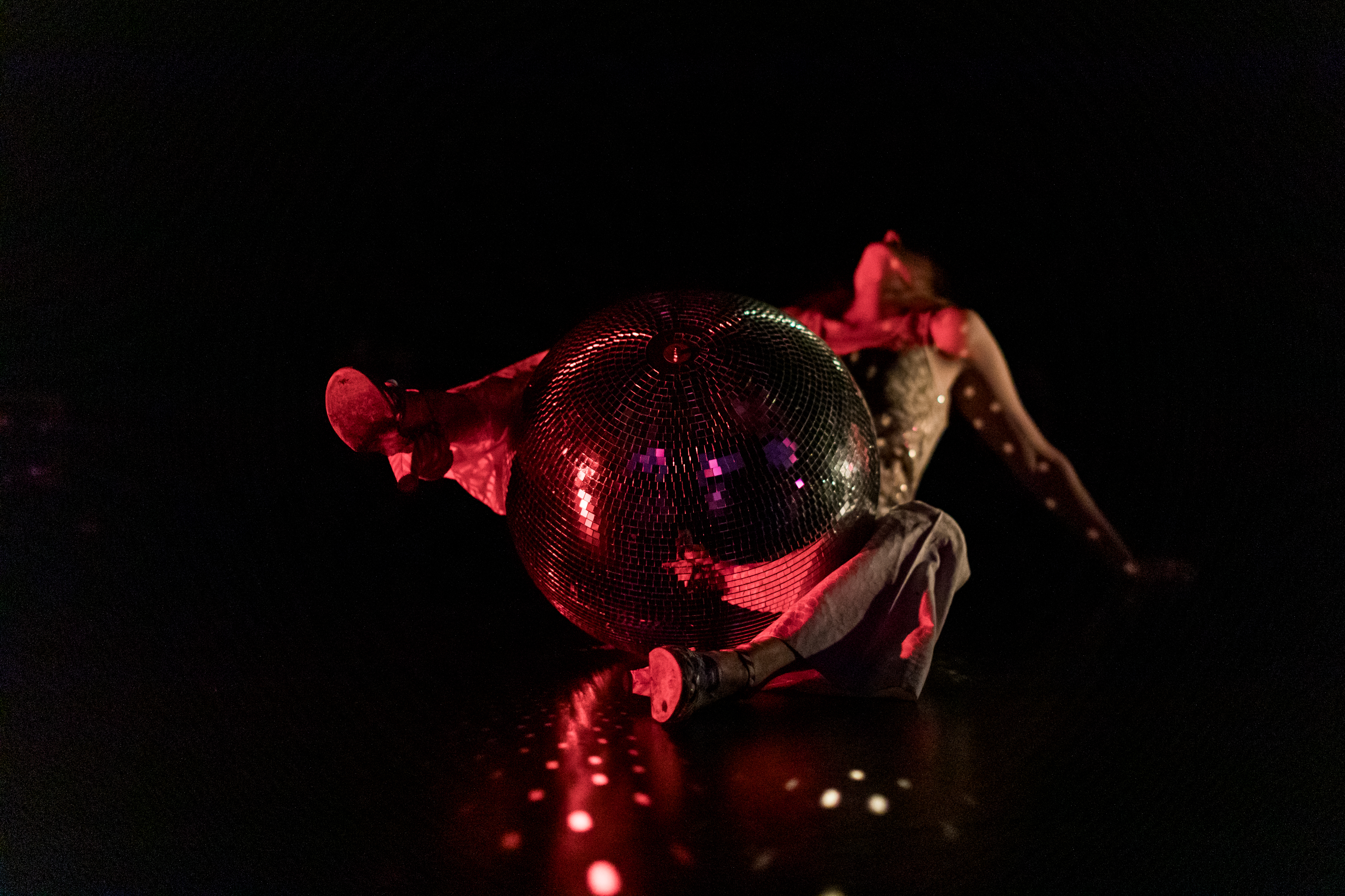 Against a dark backdrop, Peggy Piacenza straddles a disco ball which reflects spots of red light. The disco ball and Piacenza’s shoes are in focus, and her head and torso are  blurred in the background.