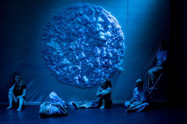In soft blue lighting, a large circle made of a cotton-like material hangs on the wall above five performers gazing to the left and sitting in a line in various relaxed poses. The rightmost performer sits on stairs.