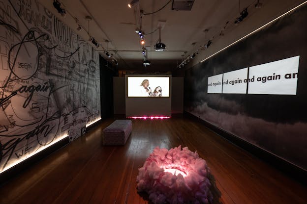 A view of an installation in a long, narrower space. The floor contains a light is surrounded by cotton candy and the far wall shows a projected image of two people with singing into microphones. The left wall is covered in the stylized written words “we begin again” in different orientations and sizes. On the right wall, the words “and again” are projected over a black and white image of clouds.