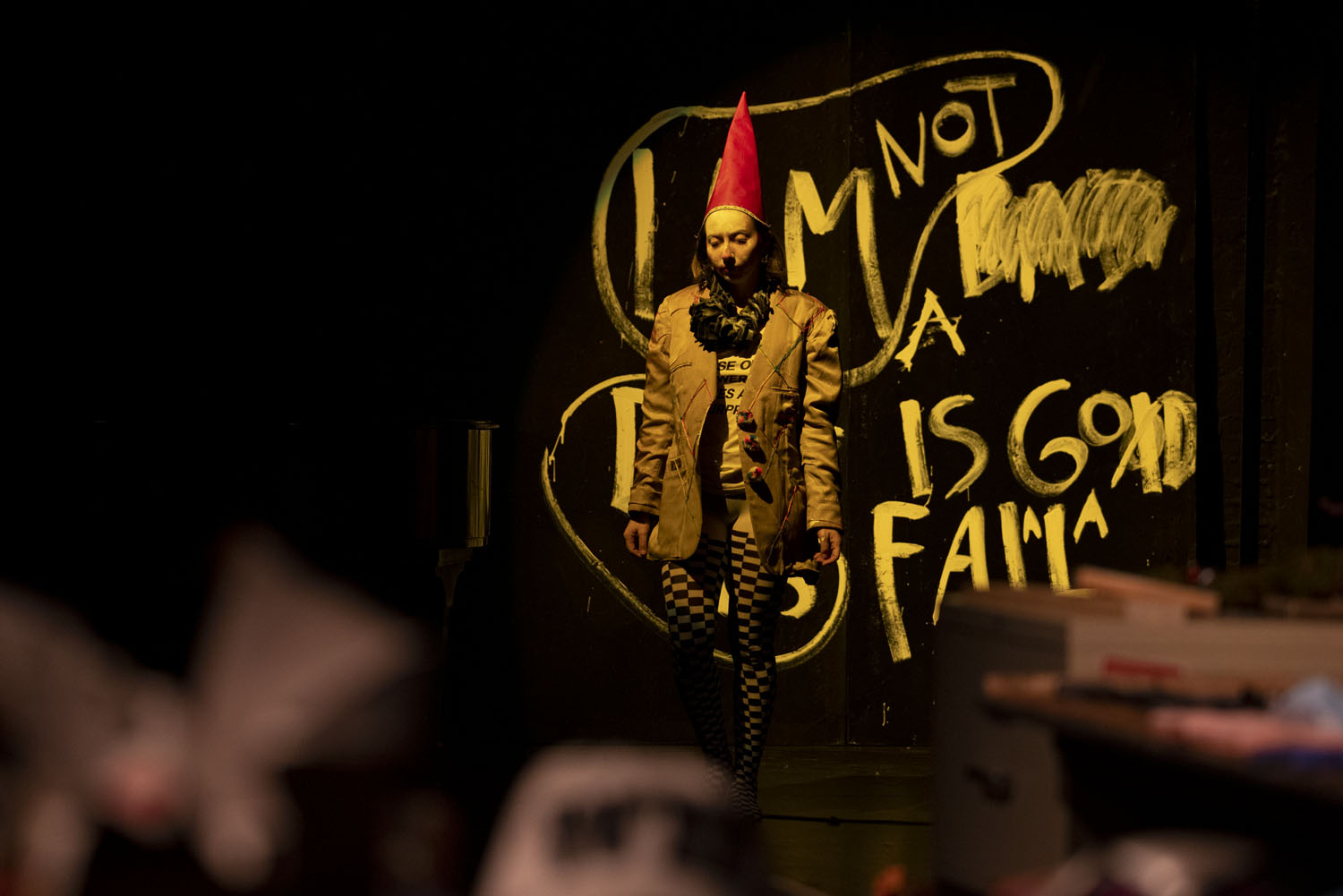 Alex Tatarsky stands center stage in a dimly lit performance space, wearing a red pointed hat, a brown blazer, and checkered tights. Behind them, a black wall is covered in large white hand-draw text that reads 