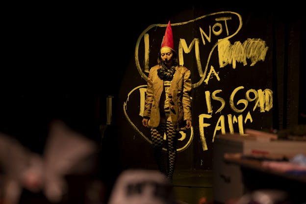 Alex Tatarsky stands center stage in a dimly lit performance space, wearing a red pointed hat, a brown blazer, and checkered tights. Behind them, a black wall is covered in large white hand-draw text that reads