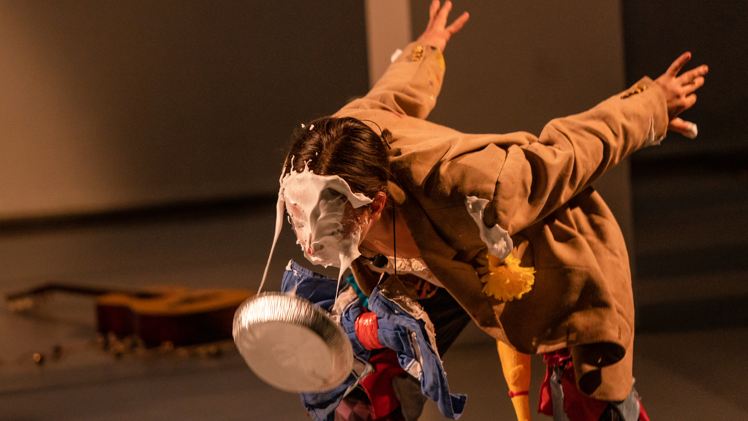 Alex Tatarsky bends forward with arms extended back and their face covered in dripping whipped cream from a pie tin. They wear a tattered brown blazer with a yellow flower pinned to the lapel and a large blue bowtie. A guitar and small scattered objects are visible on the floor in the background.