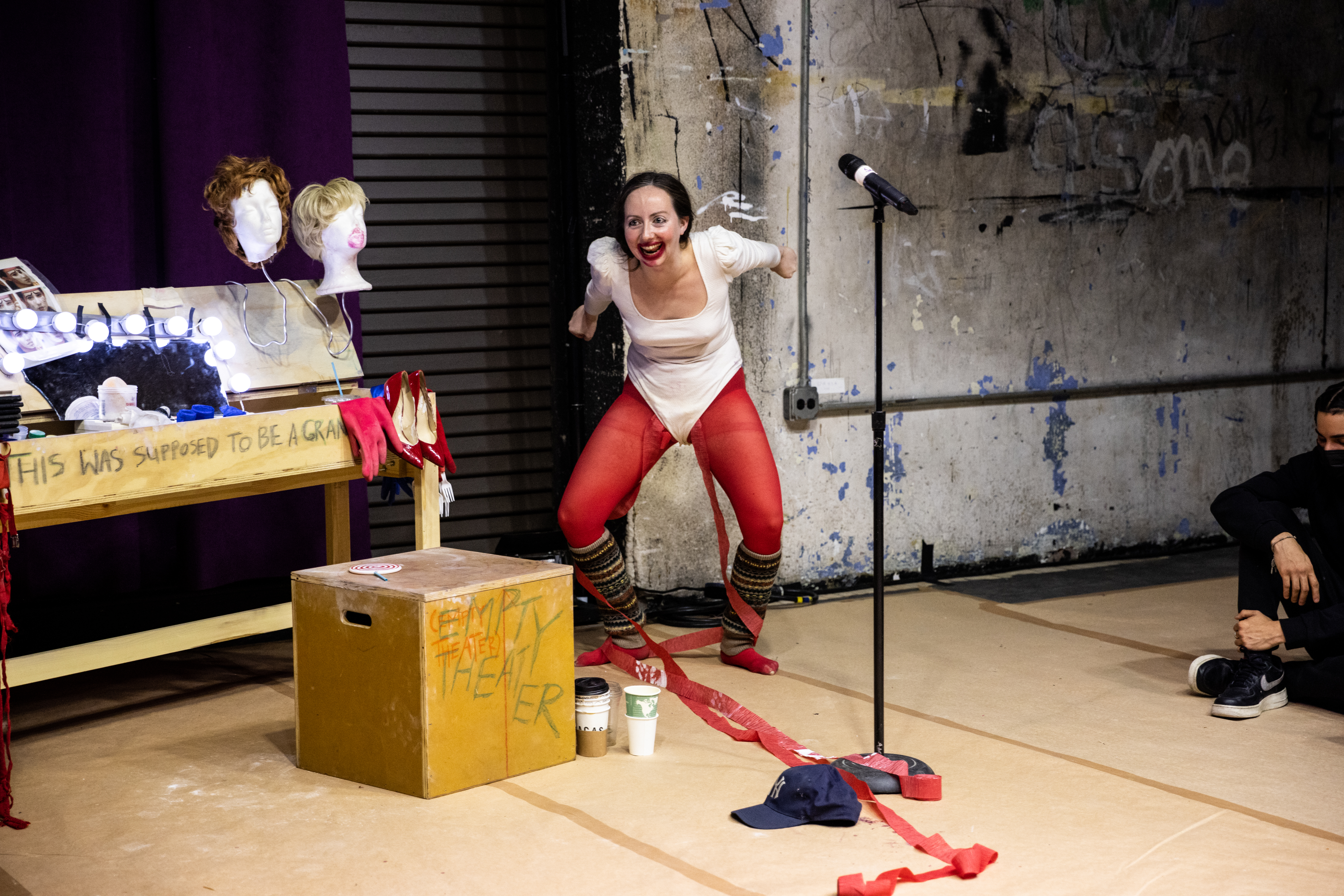 Alex Tatarsky crouches on a makeshift stage, leaning forward with arms bent back at the elbows and smiling widely, with lipstick applied on and around their mouth . They wear a white leotard with red tights and striped leg warmers. The theater set includes a wooden table with wigs, red high heels, and other props. The words 