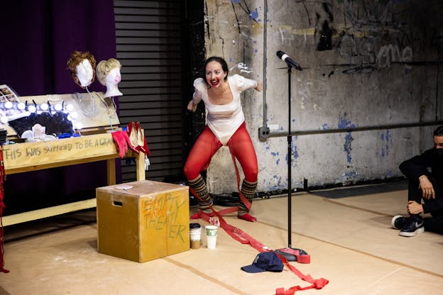 Alex Tatarsky crouches on a makeshift stage, leaning forward with arms bent back at the elbows and smiling widely, with lipstick applied on and around their mouth . They wear a white leotard with red tights and striped leg warmers. The theater set includes a wooden table with wigs, red high heels, and other props. The words