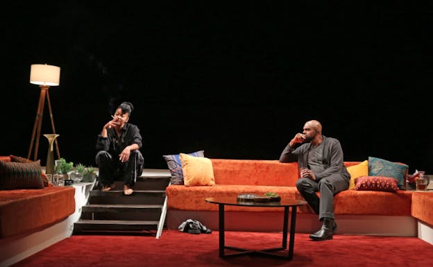Against a black background, performers Crystal Dickinson and Carl Clemons-Hopkins sit on stairs and an orange couch respectively. Dickinson wears a black shirt and pants and smokes a cigarette. Clemons-Hopkins wears a grey shirt, sweater, and pants and black boots, and looks over at Dickinson. On the ground in front of the performers, a glass coffee table sits on a red rug and the room is filled with throw pillows, house plants, and a lamp.