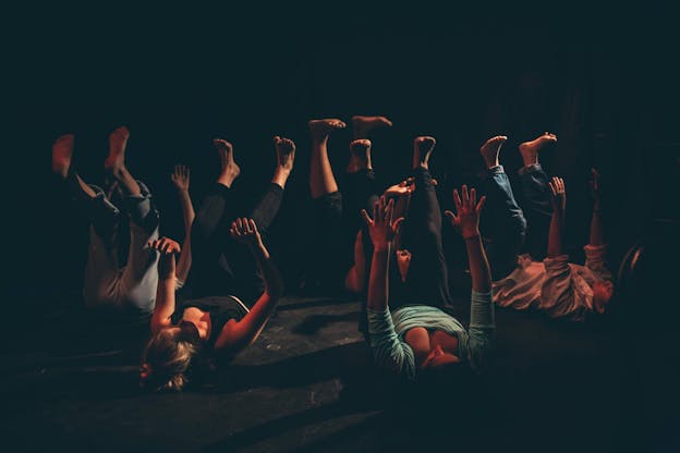 Five performers, Sarah Traisman, Genevieve Beaudoin, Amanda Centeno, Nikita Chaudhry, and Raquel Palmas, lie on their backs with their feet and hands reaching upwards. The performers lay close to one another in a circle-like formation, all facing different directions. The background is dark and the performers’ bodies are partially illuminated by warm lights and a soft spotlight. Their faces are obscured by shadows.