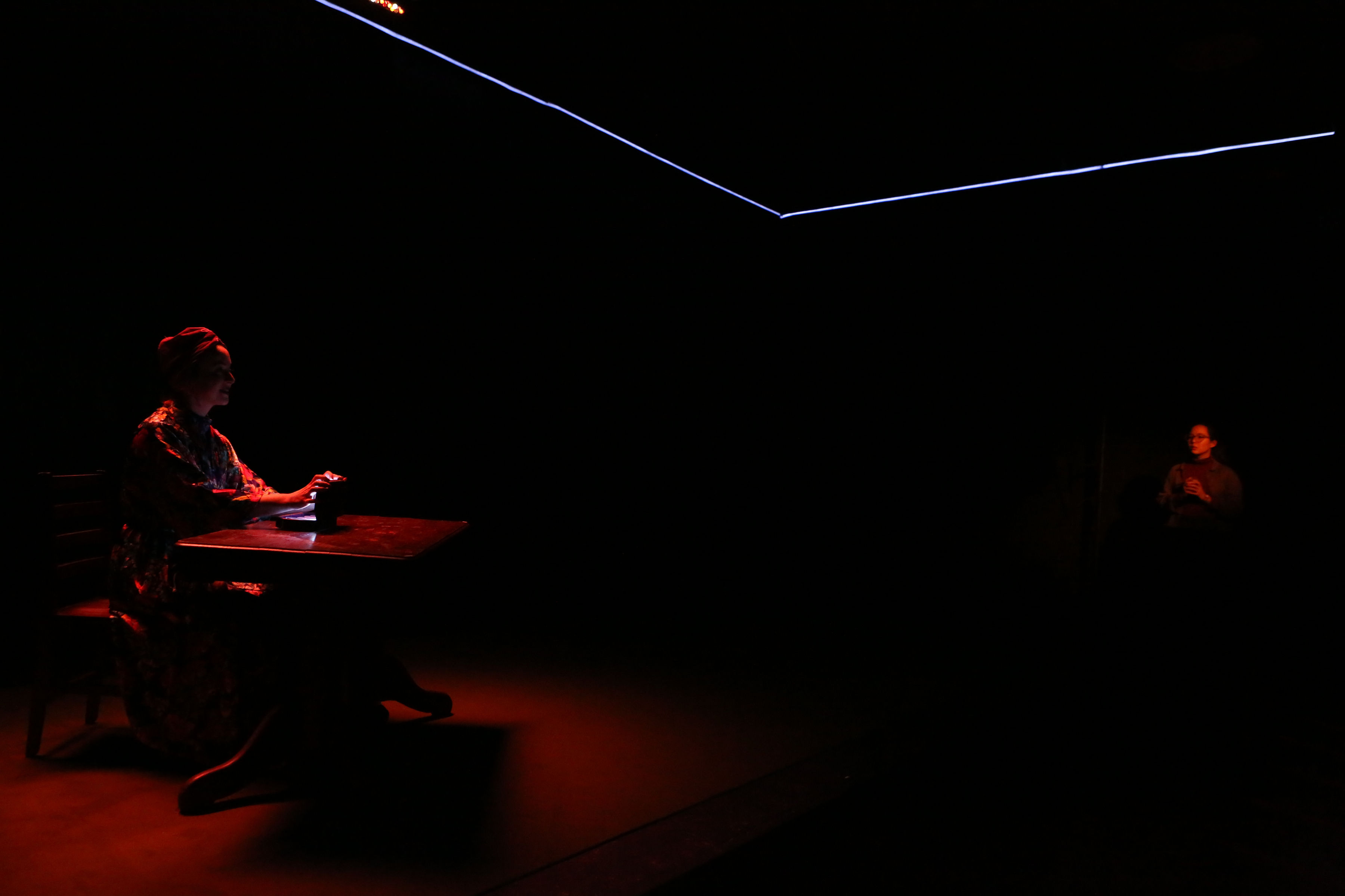 In a dark room, a performer, illuminated by a dim red light, sits behind a table and holds a glowing object. Across the image to the right, another performer, wearing glasses, is faintly visible and also illuminated by a dim red light. A blue laser-like light outlines corners of the ceiling. 