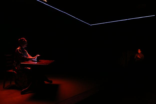 In a dark room, a performer, illuminated by a dim red light, sits behind a table and holds a glowing object. Across the image to the right, another performer, wearing glasses, is faintly visible and also illuminated by a dim red light. A blue laser-like light outlines corners of the ceiling.