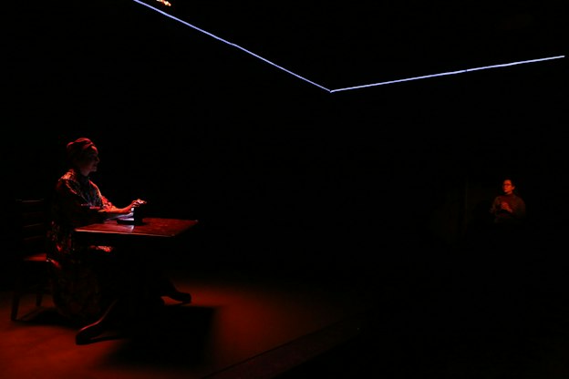 In a dark room, a performer, illuminated by a dim red light, sits behind a table and holds a glowing object. Across the image to the right, another performer, wearing glasses, is faintly visible and also illuminated by a dim red light. A blue laser-like light outlines corners of the ceiling. 