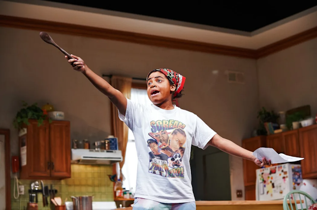 Samantha Miller stands in a kitchen with both arms extended outwards. Miller holds a paper in one hand and a wooden spoon in the other. With the wooden spoon, Miller gestures upwards. Miller wears a red bandana and a white graphic t-shirt. 