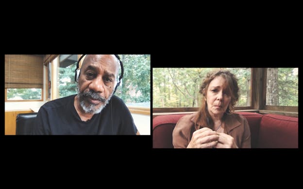 Joe Morton and Deirdre O’Connell are gazing directly into the camera in separate frames on a video conference call. Joe Morton is wearing headphones and a black shirt while sitting in front of an open window, through which trees are visible. Deirdre O’Connell, in the other frame, sits on top of a red couch and is wearing a brown collared shirt and hoop earrings. O’Connell also sits in front of an open window, through which trees are visible, and her hands are clasped together.