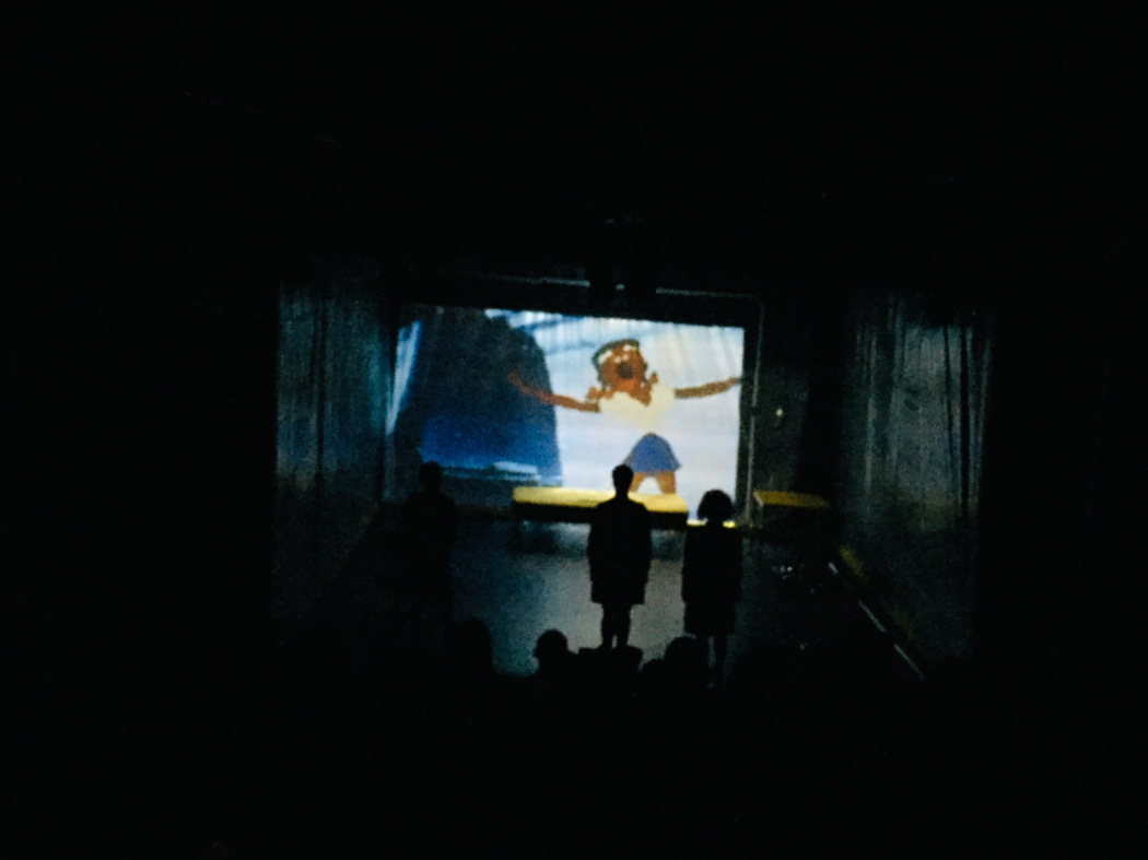 In a dark theater, an animation is projected onto a screen at the back of the stage. The figure in the animation is wearing a yellow top, blue bottoms, and their arms are outstretched. Three performers stand in silhouette in front of the screen. 