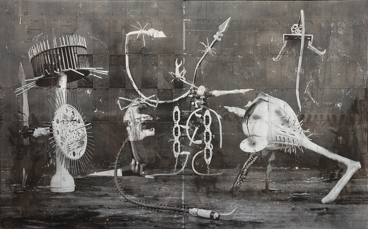 A black and white photograph shows of a group of five found-object sculptures situated arranged in front of a dark grey wall made out from of patched metal. The sculptures have are made out of tools, spikes, limbs, and blades, and disc components that make them appear in motion.