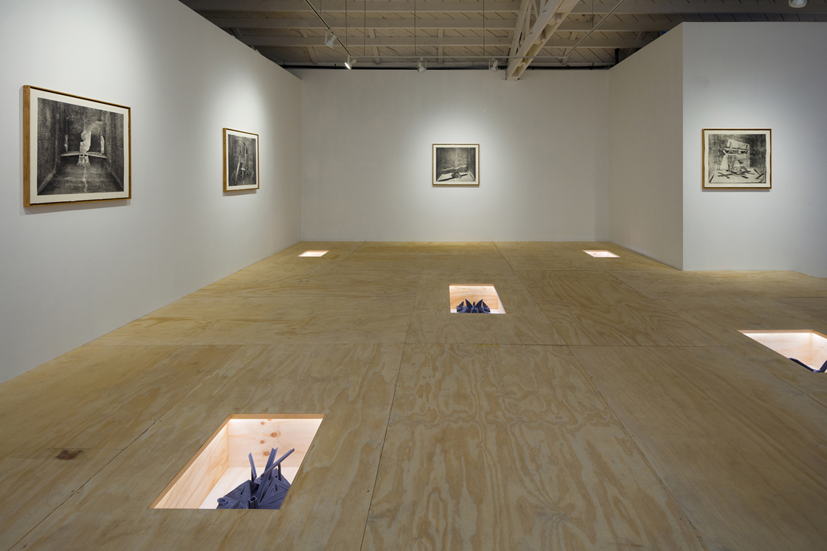 In a gallery, four black and white images hang framed on white walls. The wooden floor contains five sunken rectangular slots that house geometric sculptures. Each of the slots is illuminated so you can see the sculptures inside, which heresemble battleships, architectural models, and insects.