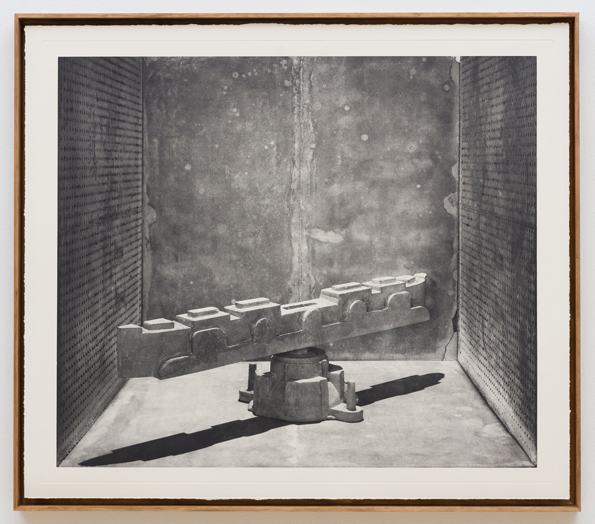  A framed black and white photograph depicts a mechanical sculpture sitting in a room. The sculpture consists of a horizontal segmented column balanced on a round base. The form resembles a piece of construction equipment and casts a long shadow across the floor. The sculpture is surrounded by speckled metal walls.