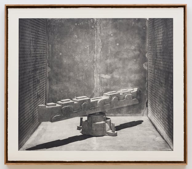 A framed black and white photograph depicts a mechanical sculpture sitting in a room. The sculpture consists of a horizontal segmented column balanced on a round base. The form resembles a piece of construction equipment and casts a long shadow across the floor. The sculpture is surrounded by speckled metal walls.